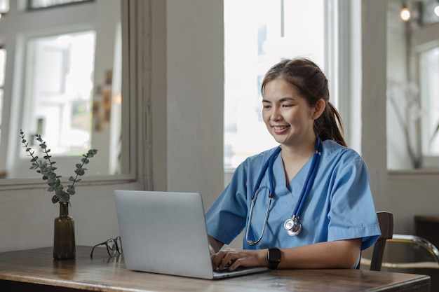 young asian lady doctor in white medical uniform with stethoscope using computer laptop talking video conference call with patient at desk in health clinic or hospital