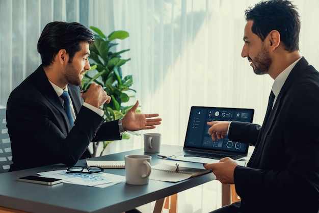 Two men sitting at a table, one of them is pointing at a screen that says'business plan '