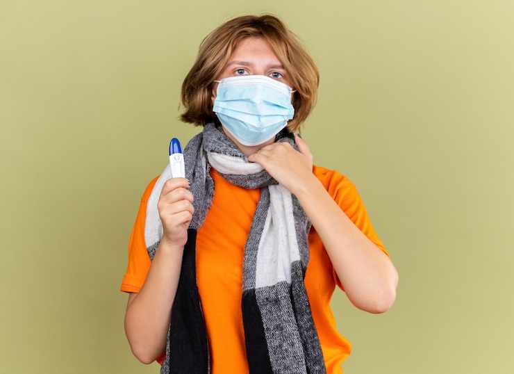 Unhealthy young woman with warm scarf around her neck wearing protective facial mask holding thermometer suffering from throat sore touching neck standing over green wall