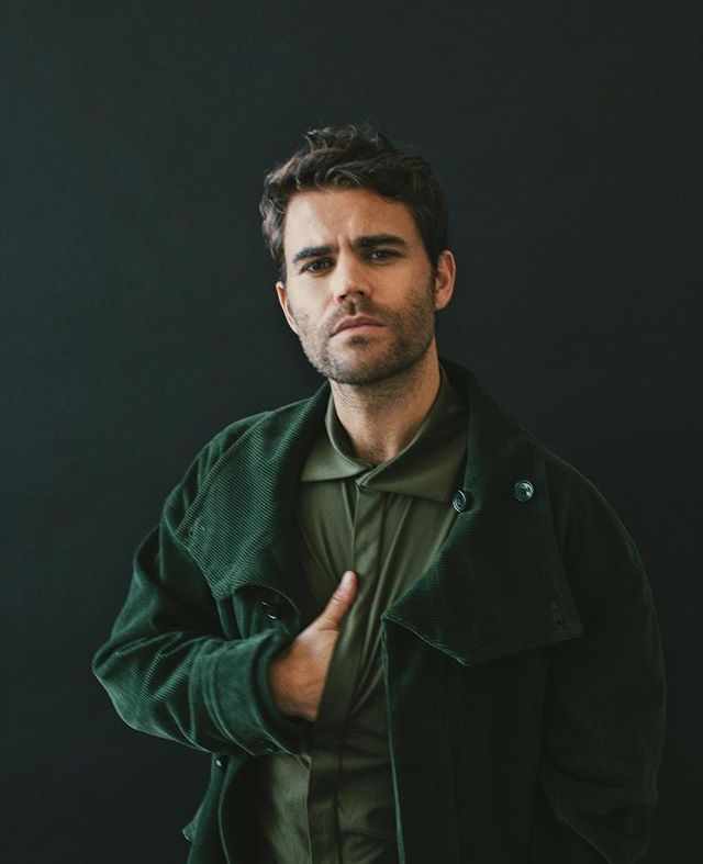 This may contain: a man in a green jacket standing with his hand on his chest and looking at the camera