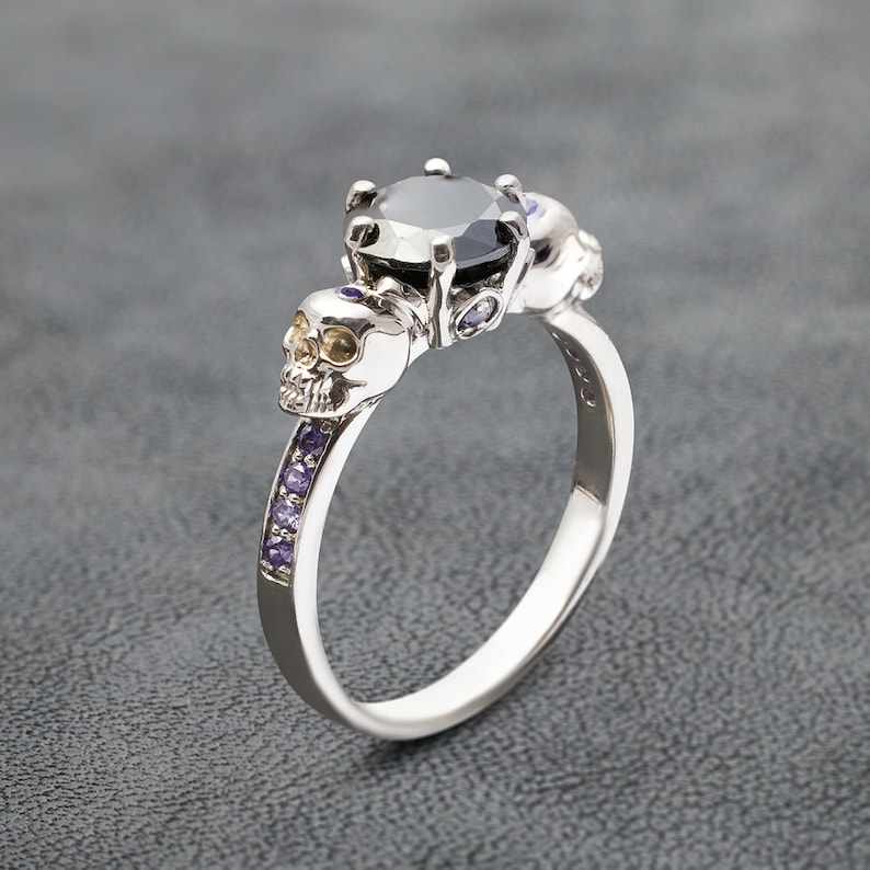 May include: A silver ring with a black gemstone and two skull accents. The ring is set with small purple gemstones.