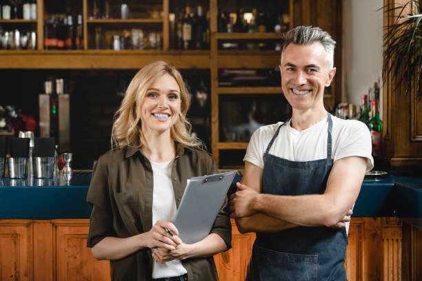 Smiling caucasian small business owner waiter barista with female auditor holding clipboard standing at the bar restaurant cafe counter checking products quality looking at the camera Smiling caucasian small business owner waiter barista with female auditor holding clipboard standing at the bar restaurant cafe counter checking products quality looking at the camera franchise business stock pictures, royalty-free photos & images