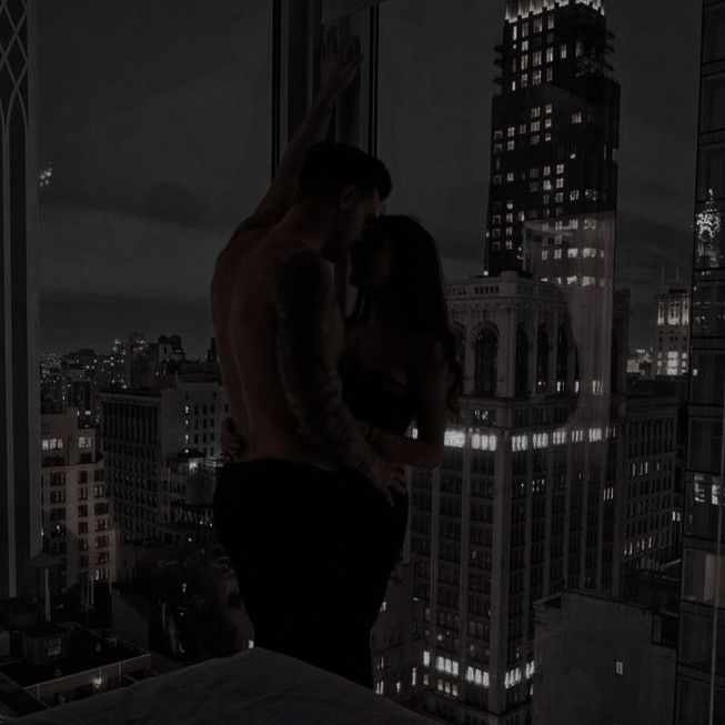 This may contain: two people are standing in front of a window overlooking the city skyline at night time